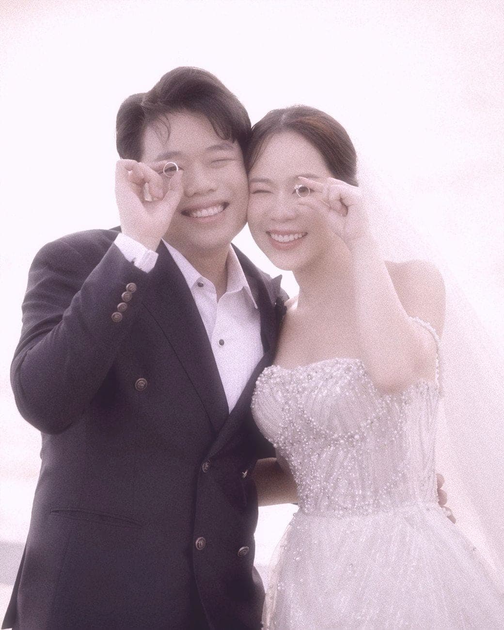 Witita and Sorratat holding their wedding rings up to one eye, by the sea